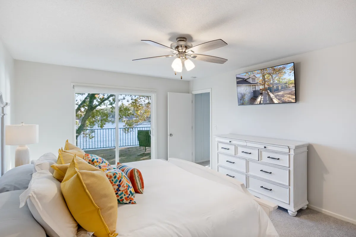 A tranquil bedroom with a bed topped with colorful pillows, a large window framing beautiful lake views, a ceiling fan overhead, and a television for relaxing evenings. Soft morning light fills the space, creating a peaceful, welcoming atmosphere.