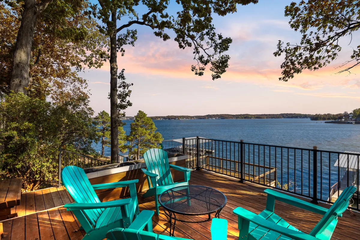 The image showcases turquoise Adirondack chairs arranged on a spacious deck overlooking tranquil lake waters, with a vivid sunset painting the sky in warm hues. The scene invites relaxation, socializing, and enjoying the breathtaking views from the comfort of the deck, with the peaceful lake and natural surroundings creating a serene atmosphere.