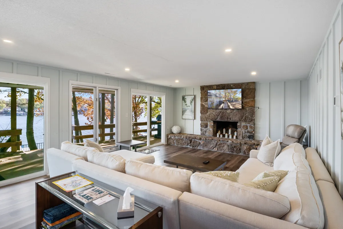 This image features a large sectional sofa facing a striking stone fireplace, with expansive windows offering stunning views of the lake. The arrangement is perfect for family gatherings or cozy movie nights, with plenty of natural light and a welcoming atmosphere.