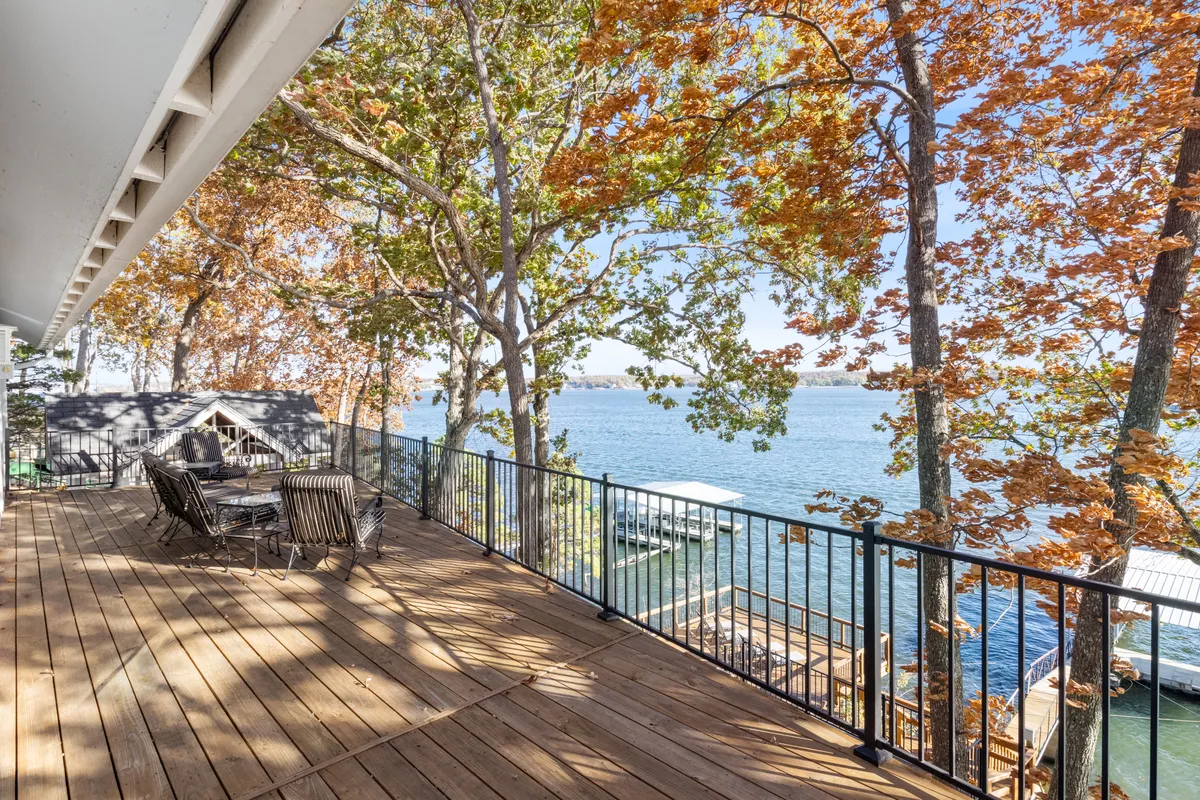 A spacious wooden deck overlooks a tranquil lake framed by vibrant autumn foliage. Outdoor seating is arranged to invite relaxation or al fresco dining, with the glistening water just steps away and the colors of fall reflected in the lake.